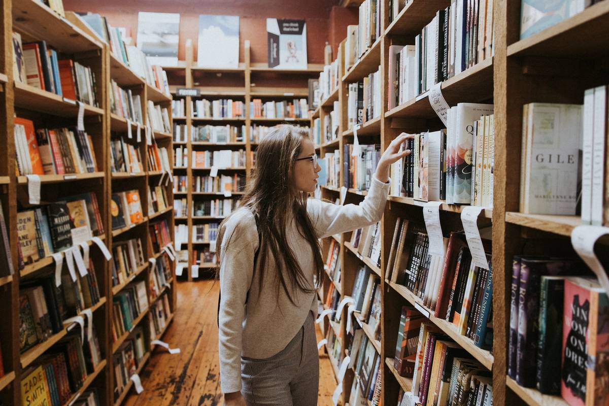 Kobieta w okularach przegląda książki znajdujące się na regale w bibliotece.