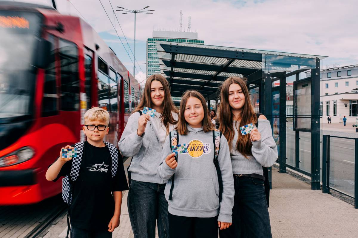 Trzy dziewczynki i chłopiec stoją na przystanku tramwajowym i trzymają Kartę Mieszkańca.